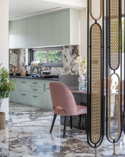 Kitchen with sage cabinetry and marble backsplash seen through a cane-and-brass room divider, pink chair beyond