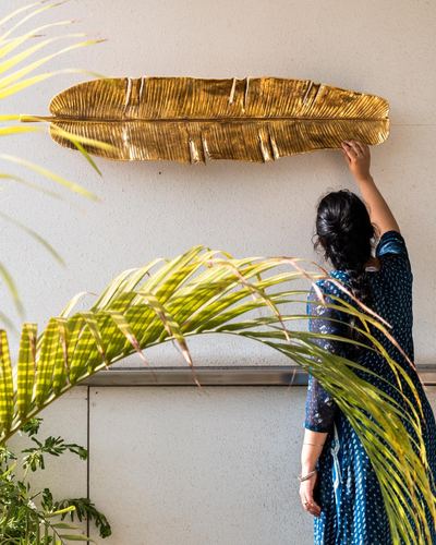 Woman in blue sari adjusting a large gold leaf-shaped wall sculpture beside palm fronds