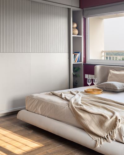 Sunlit bedroom corner with fluted wardrobe, open bookshelf, and upholstered bed with knit throw.