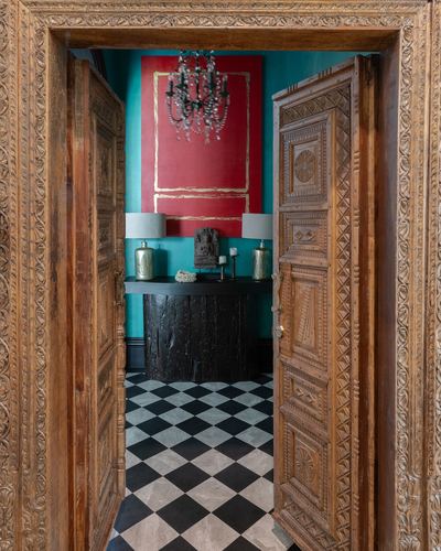 View through a heavily carved wooden double door into a teal foyer with checkerboard floor and red art panel