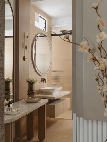 Beige stone bathroom with round mirror, vessel sink, and fluted glass partition seen through doorway