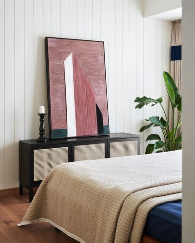 Bedroom corner with cane credenza, leaning abstract painting, candle, and banana plant beside bed