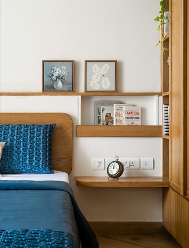 Bedroom corner with wood headboard, blue quilted pillow, floating shelves with books and framed art