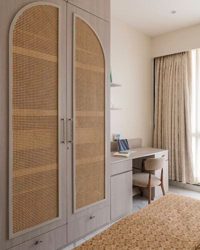 Bedroom corner with arched cane wardrobe doors, wooden desk, chair, and beige curtains