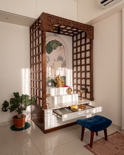 Wooden lattice-framed pooja unit with marble steps, brass idol, and blue velvet stool in sunlit corner