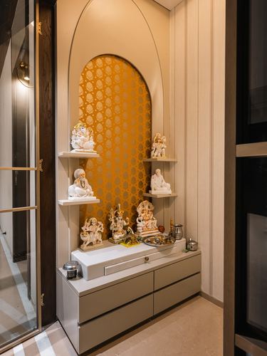 Arched pooja niche with mustard patterned wall, floating shelves holding idols, above a storage cabinet