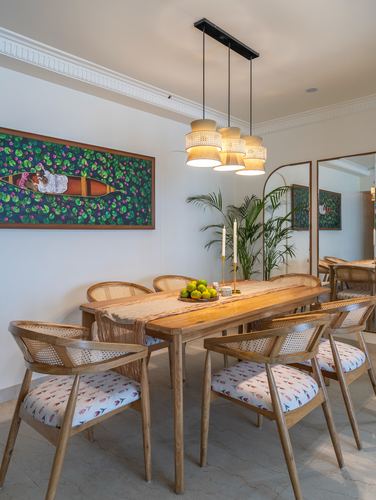 Wooden dining table with cane chairs beneath a cluster pendant light and framed folk painting