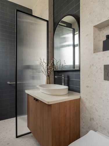 Bathroom vanity with wood cabinetry, vessel sink, arched mirror and fluted glass shower partition