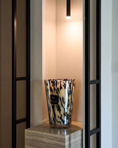Speckled glass vase on travertine pedestal lit by a pendant, framed by black glass partitions in a Dubai villa entry.