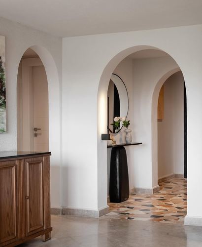 Hallway with three plastered arches, a wooden cabinet, round mirror, and patterned terrazzo floor beyond