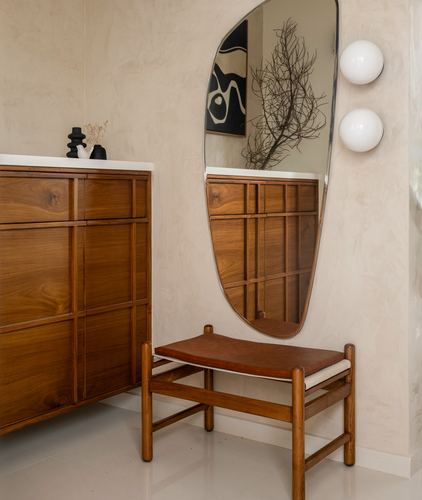 Wooden cabinet, organic mirror, and leather bench against textured plaster wall with sconces