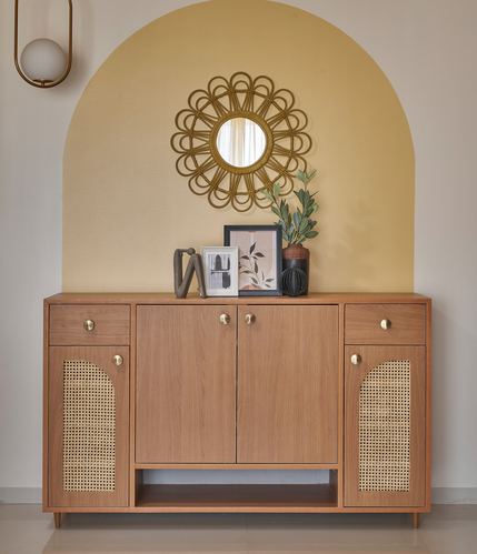 Wooden cane-front sideboard beneath arched mustard wall with sunburst mirror and decor