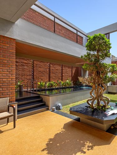Brick jaali courtyard with raised pool, cane chair, and sculptural tree on stone platform.