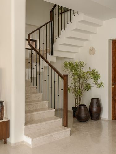 Curved staircase with teak handrail and black balusters beside foyer plants and urns