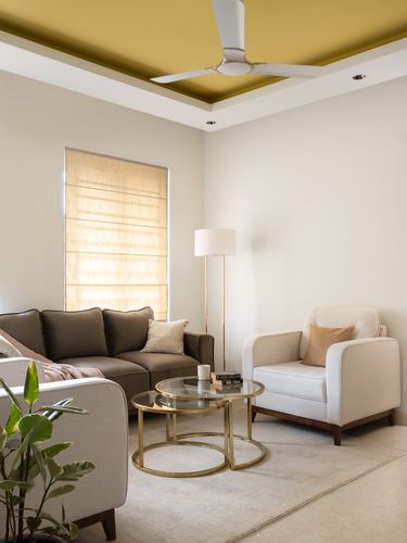 Beige living room with mustard tray ceiling, brown sofa, cream armchairs, and glass nesting tables