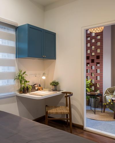 Bedroom corner with teal wall cabinet, floating desk, cane chair, and view to balcony with jali screen