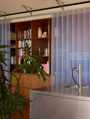 Kitchen island with steel counter, sheer curtain, and wooden bookshelf behind foliage