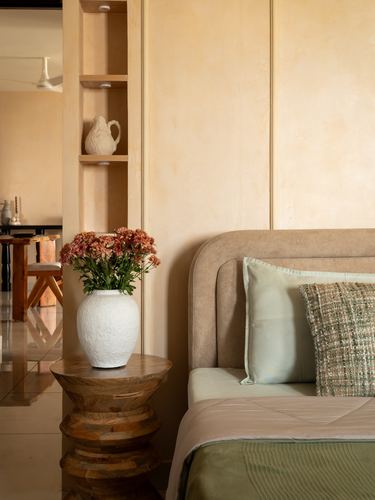 Bedroom corner with upholstered headboard, wooden side table, white vase and flowers against lime-plaster wall