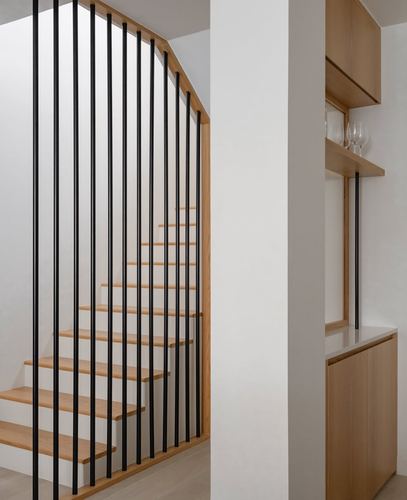 Staircase behind vertical black metal rods, wood treads, white walls, wood cabinet with glassware nearby