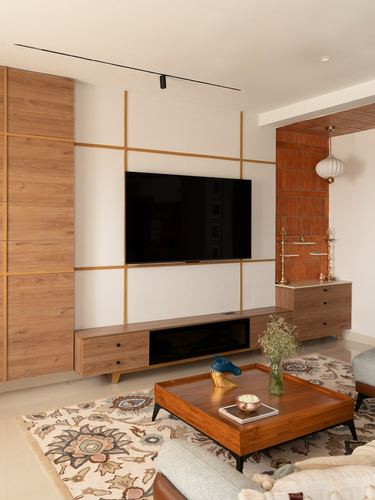 Living room with brass-grid wood TV wall, terracotta pooja niche, and wooden coffee table on patterned rug