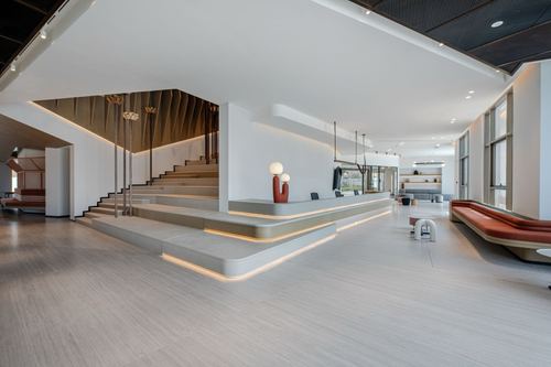 Large minimalist office lobby with backlit staircase, curved reception desk, and warm stone flooring