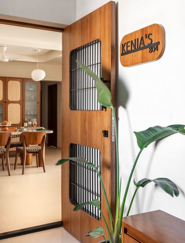 Wooden entry door with black metal grille panels beside a nameplate, plant, and view into dining room