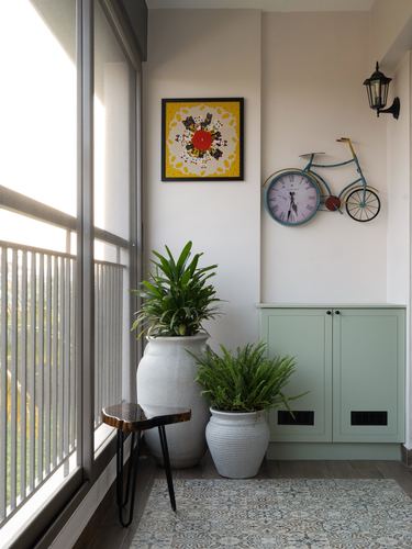 Balcony corner with plants, patterned rug, sage cabinet, and bicycle-shaped wall clock beside textile art