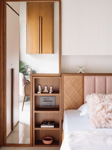 Bedroom nook with wooden shelving, cane headboard, and pink fluted wall panel beside a mirrored passage