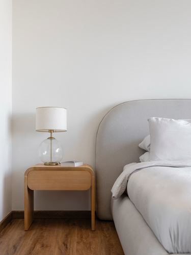 Wooden nightstand with glass lamp beside a grey curved upholstered headboard and bed