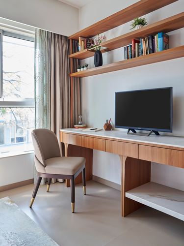 Study nook with wood desk, wall-mounted TV, floating bookshelves and a beige chair by a window