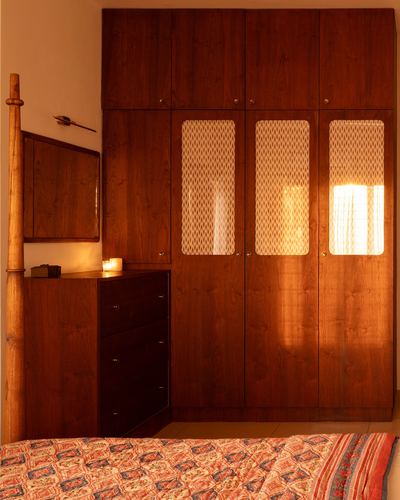Warm-lit wooden wardrobe and chest of drawers in a Kochi bedroom with patterned bedspread in foreground