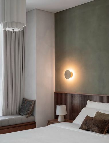 Bedroom corner with sage plaster wall, fluted wood paneling, glowing wall sconce, and window seat