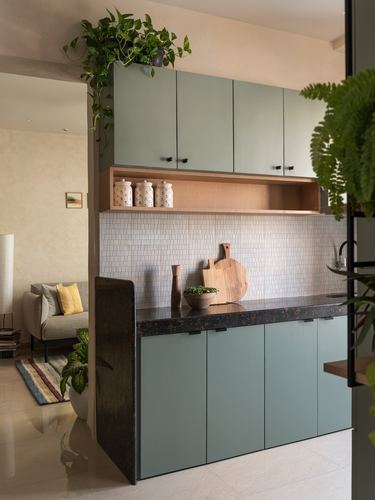 Sage green kitchen with textured stone counter, wood shelf, and tiled backsplash seen through archway