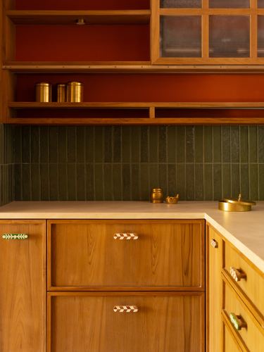 Corner of wooden kitchen cabinetry with green tile backsplash, brass canisters, and terracotta wall behind open shelving
