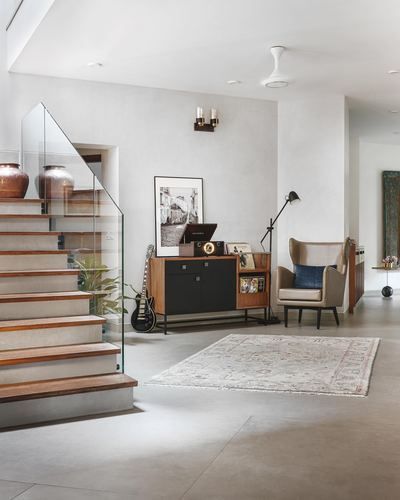 Staircase with glass railing beside a mid-century console, armchair and rug in a light-filled foyer