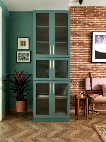 Tall forest-green glass cabinet beside exposed brick wall in a styled living corner