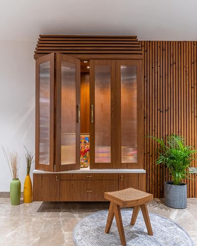 Wooden pooja cabinet with fluted glass doors lit from within, beside a reeded wood wall and cane stool