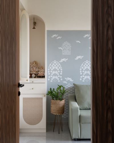 View through a fluted wood door into a pooja cabinet and living room with bird-patterned blue wallpaper