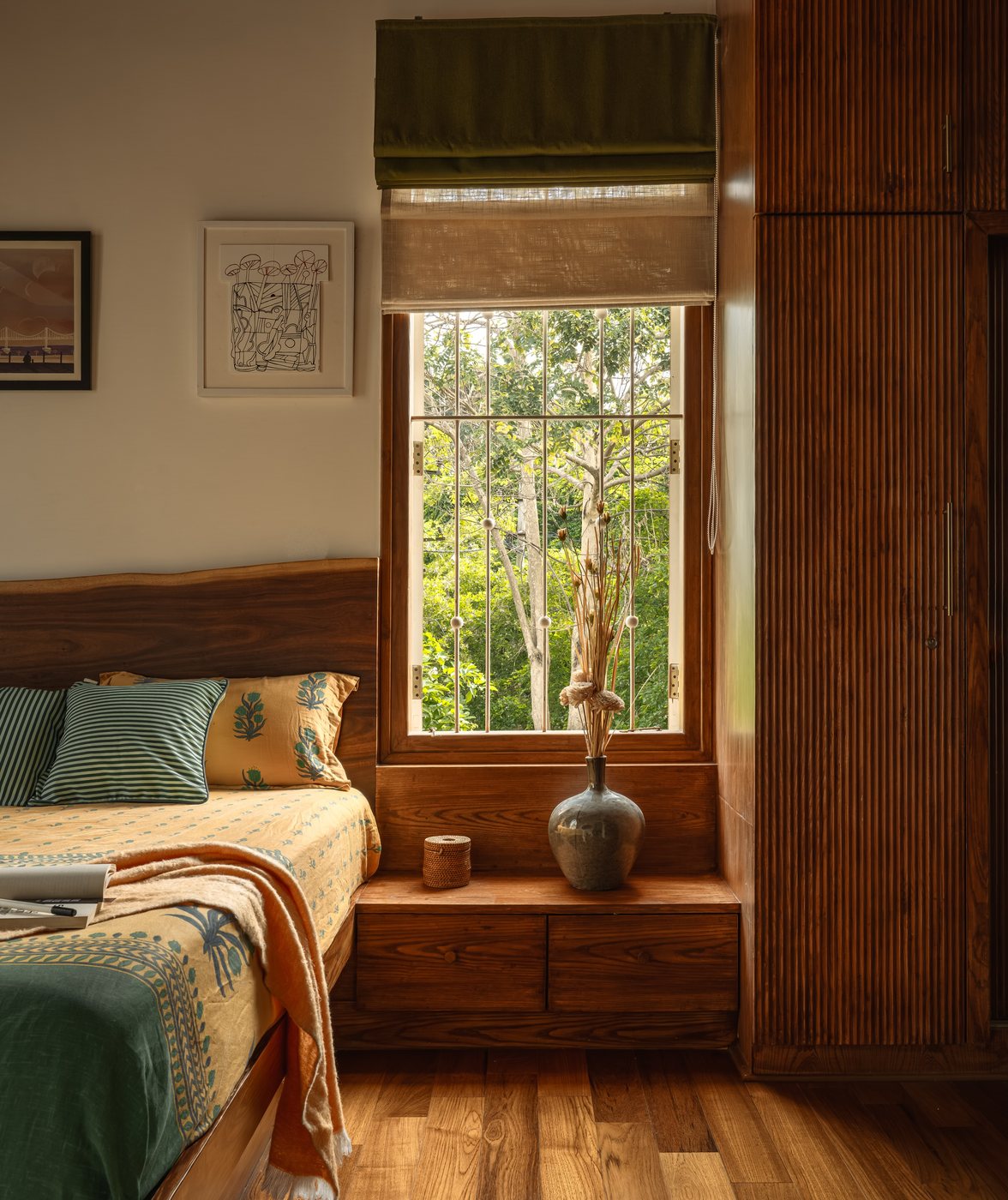 Wooden bed beside a window seat with vase, overlooking green trees, warm afternoon light