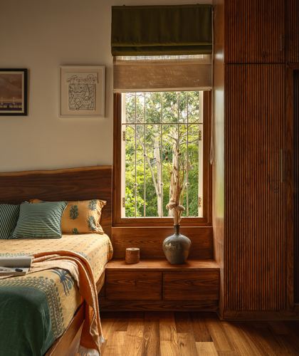 Wooden bed beside a window seat with vase, overlooking green trees, warm afternoon light