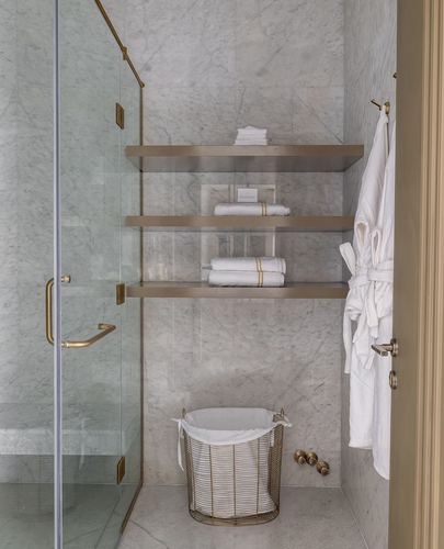 Marble bathroom corner with glass shower, brass shelves holding towels, and a robe hung on a hook