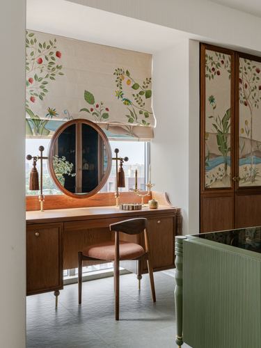 Dressing table with round mirror beneath a floral roman blind, wooden chair, and painted cabinet nearby