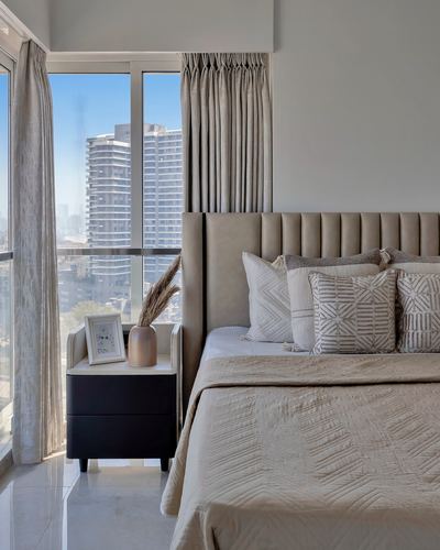 Beige bedroom with tufted headboard, textured pillows, dark nightstand and city skyline view through curtained window