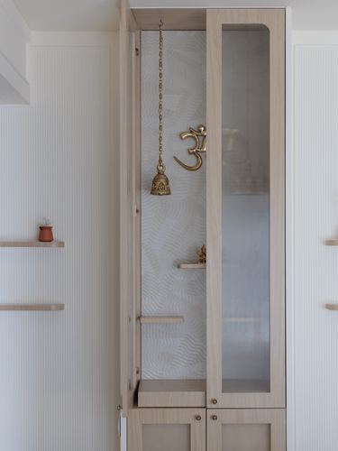 Wooden pooja cabinet with brass om symbol and hanging bell against textured wallpaper