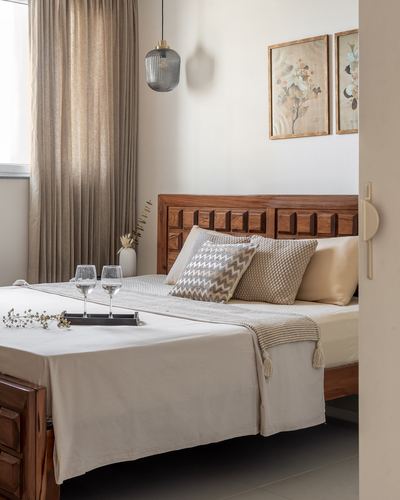 Bedroom with carved wood headboard, neutral linens, framed botanical prints and pendant lights seen through doorway