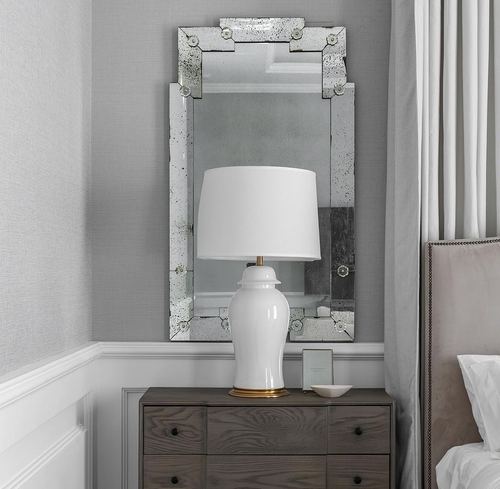 Venetian mirror above wood dresser with white ginger jar lamp beside upholstered bed