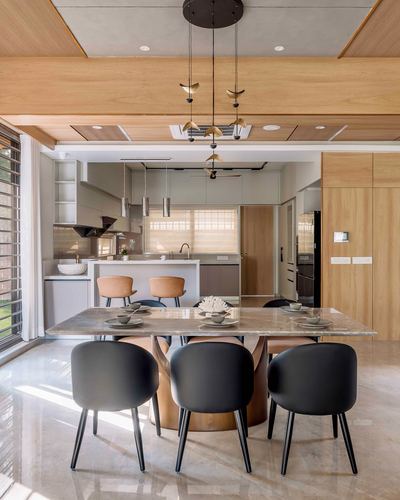 Open dining and kitchen area with marble table, black chairs, and brass leaf pendant lights under a wood-paneled ceiling
