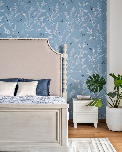 Bedroom corner with upholstered bed, carved white bedpost, botanical blue wallpaper, and potted monstera plant