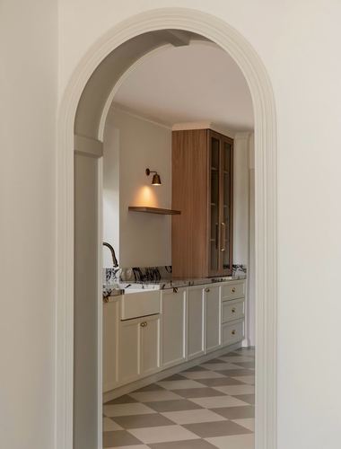 Arched doorway framing a white kitchen with marble counters, wood cabinet, and checkerboard floor