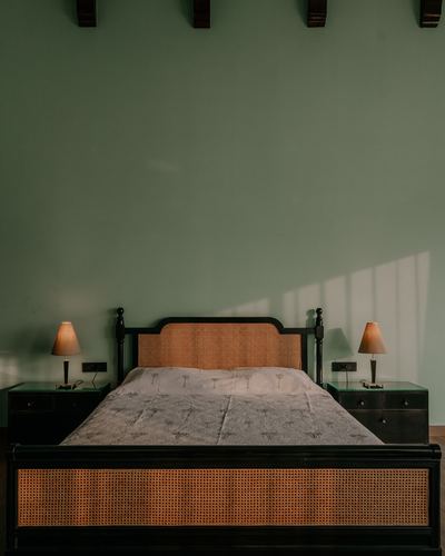 Dark wood bed with cane headboard and footboard against a sage green wall, sunlight streaking across